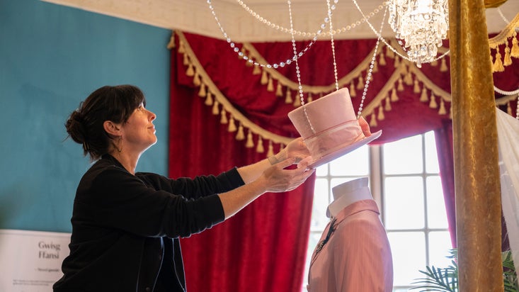 Exhibition designer places a pink top hat onto a mannequin, finishing off the costume display.
